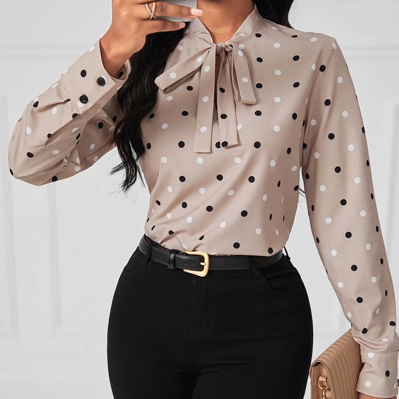 Woman wearing beige polka dot blouse with bow tie collar and black high-waisted pants, accessorized with black belt and beige handbag.