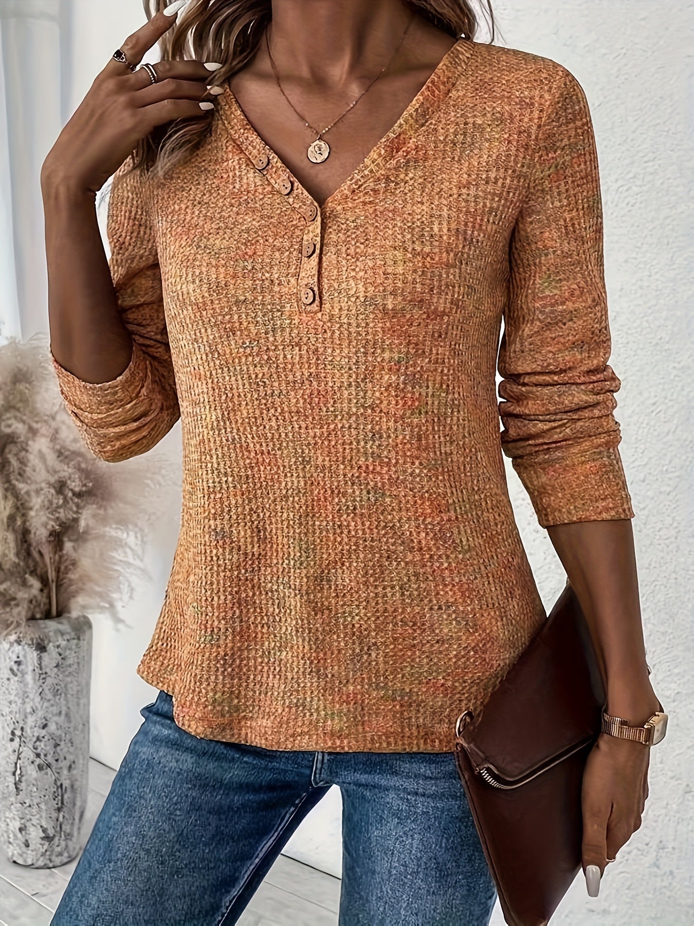 Woman wearing an orange textured long sleeve shirt with jeans, holding a brown clutch purse in a stylish indoor setting