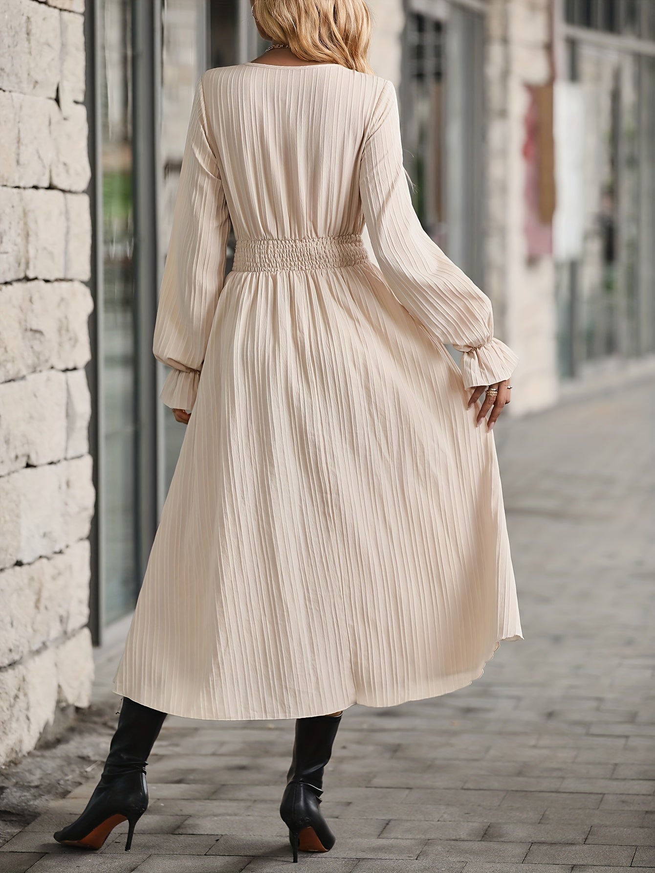 Woman wearing a long-sleeved, pleated beige dress and black ankle boots walking on a cobblestone street