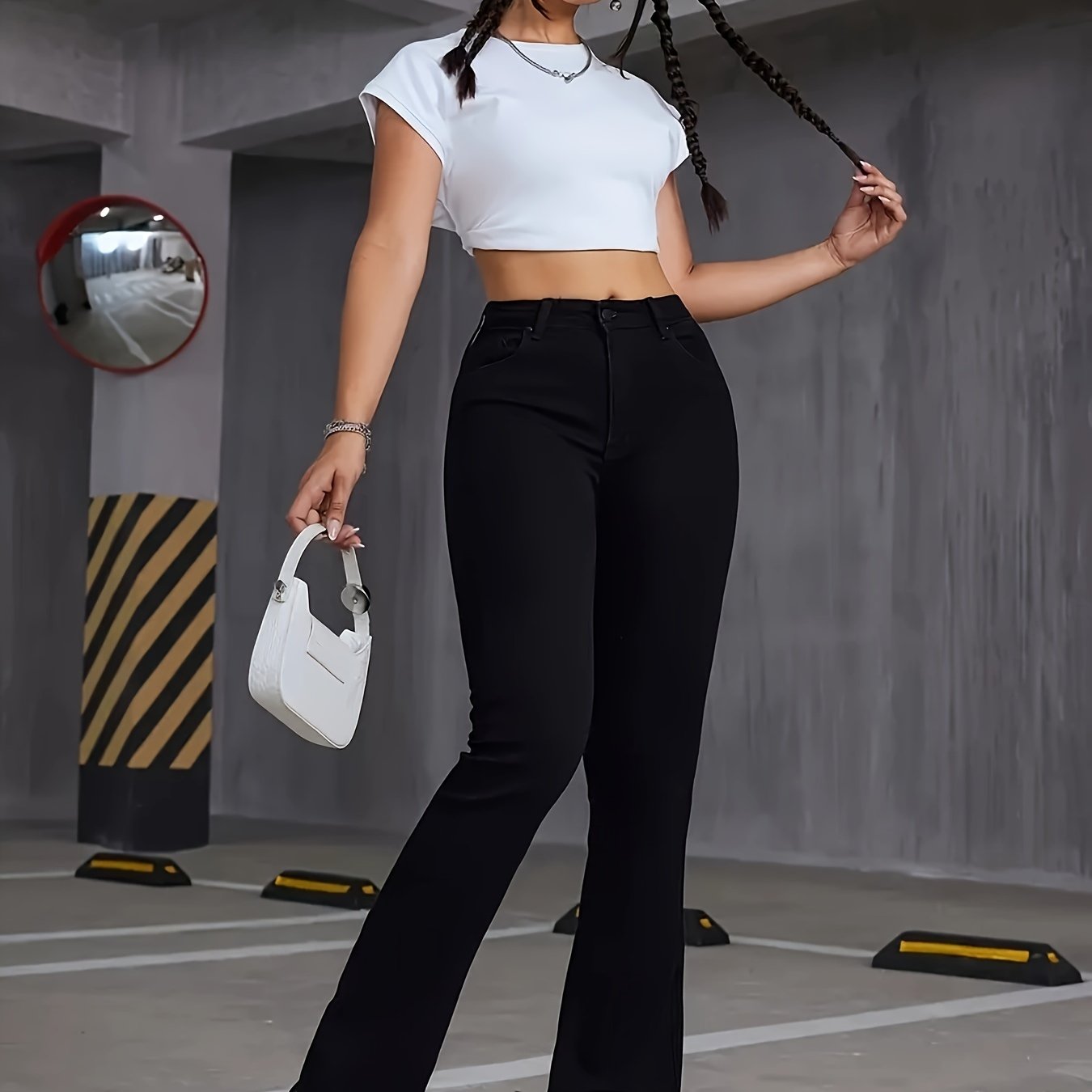 Woman in black flare leg jeans and white crop top holding a white purse in a modern, industrial setting.