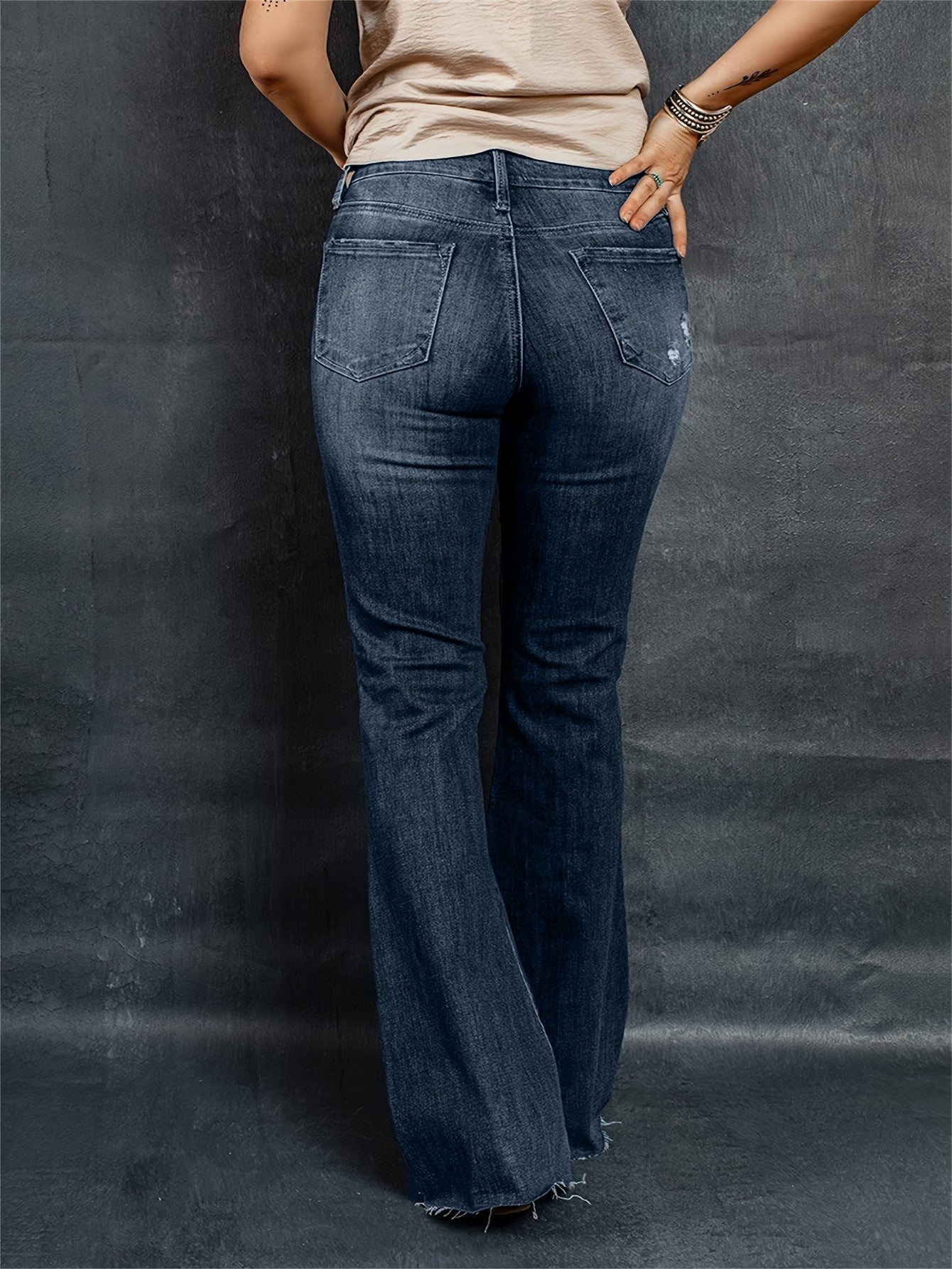 Woman wearing stylish plus-size flared leg jeans with washed ripped raw trim and button fly, showcasing back view against dark background.